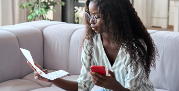 Young smart serious African black businesswoman in glasses sitting on sofa reading bank loan legal financial documents using cell mobile phone at modern home office. Distant remote work concept.