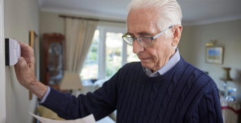 Worried Senior Man With Bill Turning Down Central Heating Thermostat At Home In Energy Crisis